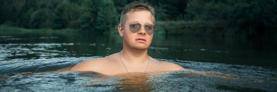 Man with sunglasses enjoys a refreshing swim in a river surrounded by lush greenery.